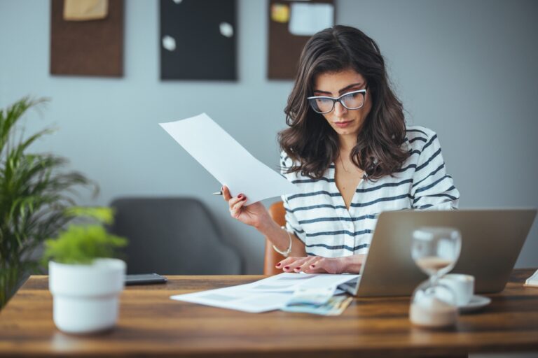 Woman in glasses working at desk with laptop, holding and reading documents.