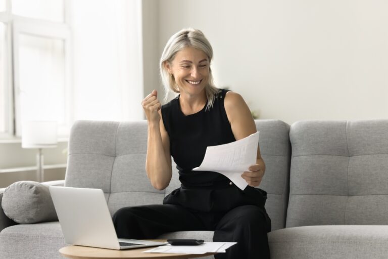 Smiling woman celebrating success while reading paper on sofa with laptop open.