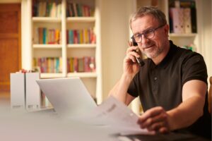 A man reviews house sale documents while speaking on the phone with his estate agent, considering whether to accept an offer on his property