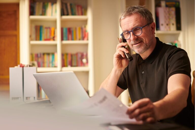 A man reviews house sale documents while speaking on the phone with his estate agent, considering whether to accept an offer on his property