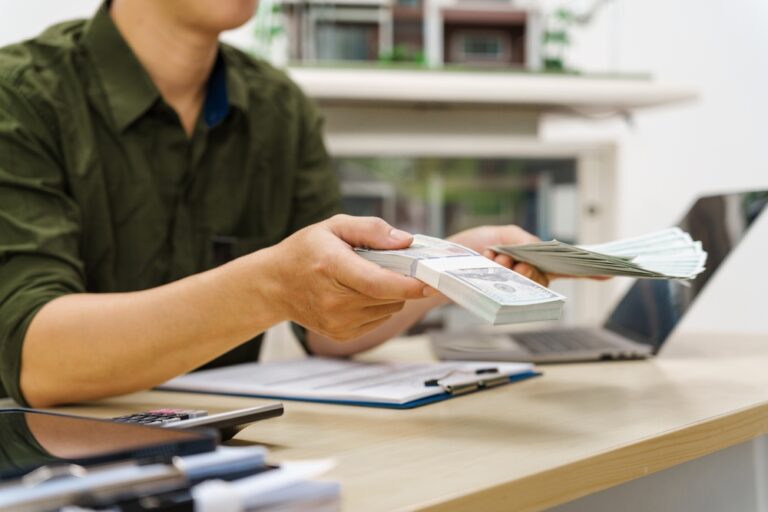 Man handing over cash bundles at a desk with laptop and documents
