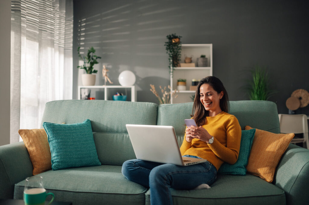 A woman relaxing on a sofa using her laptop and smartphone in a modern home, representing comfortable living in Rainhill (L35).