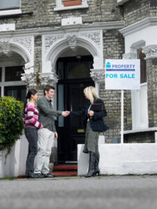A real estate agent shakes hands with a man while his partner stands beside him outside a brick house with a property-for-sale sign.
